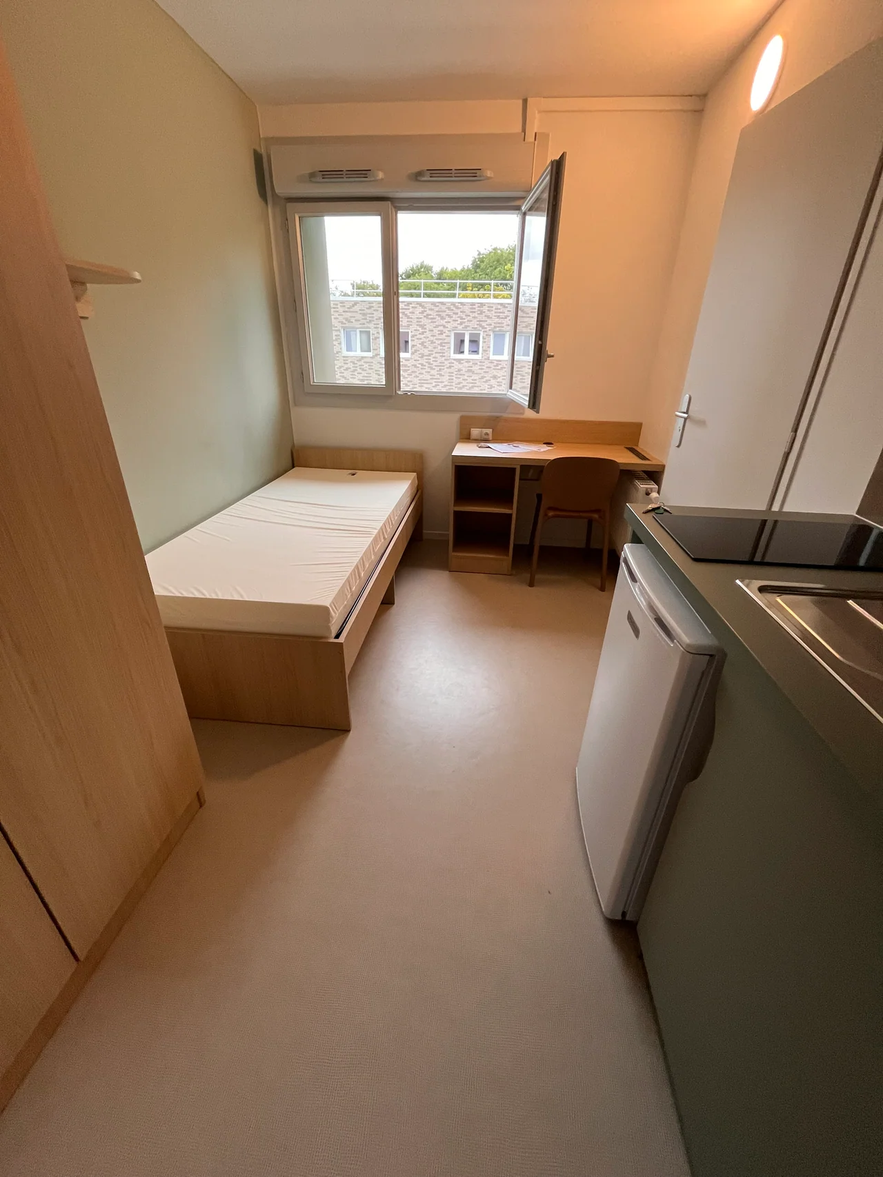 Vue d’ensemble d’une studette du bâtiment J avec kitchenette, armoire, bureau, lit, fenêtre et accès à la salle d’eau